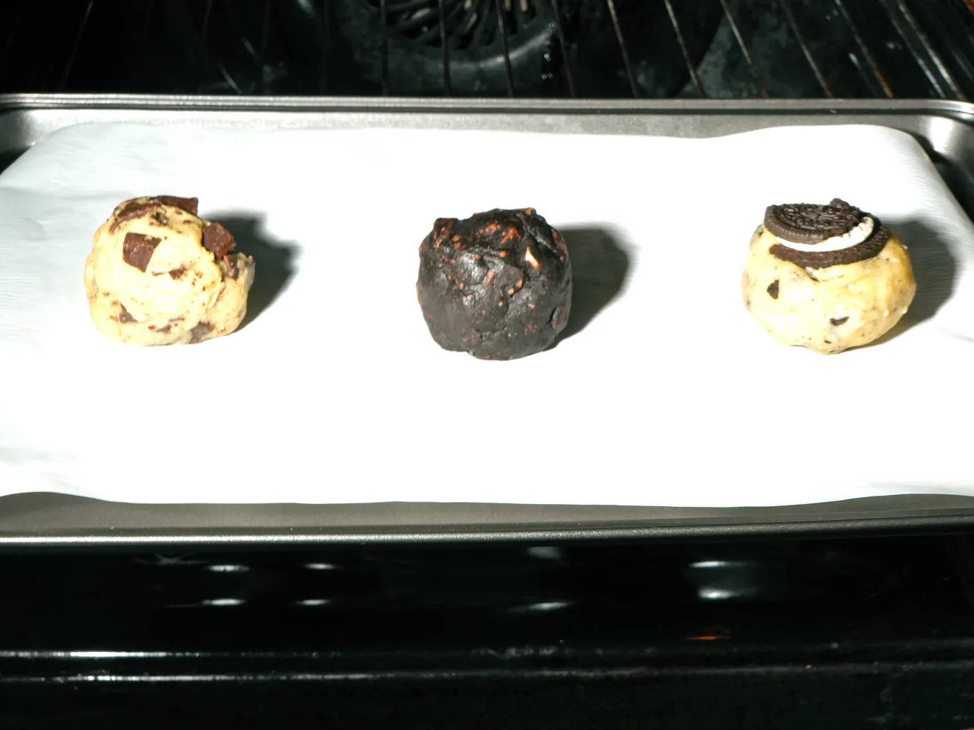 Tres bolitas de masa, una de cada sabor, sobre papel manteca en una bandeja, entrando al horno